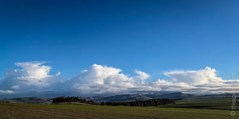 A picture across arable grassland of distant hills, with a blue sky and fluffy white clouds.